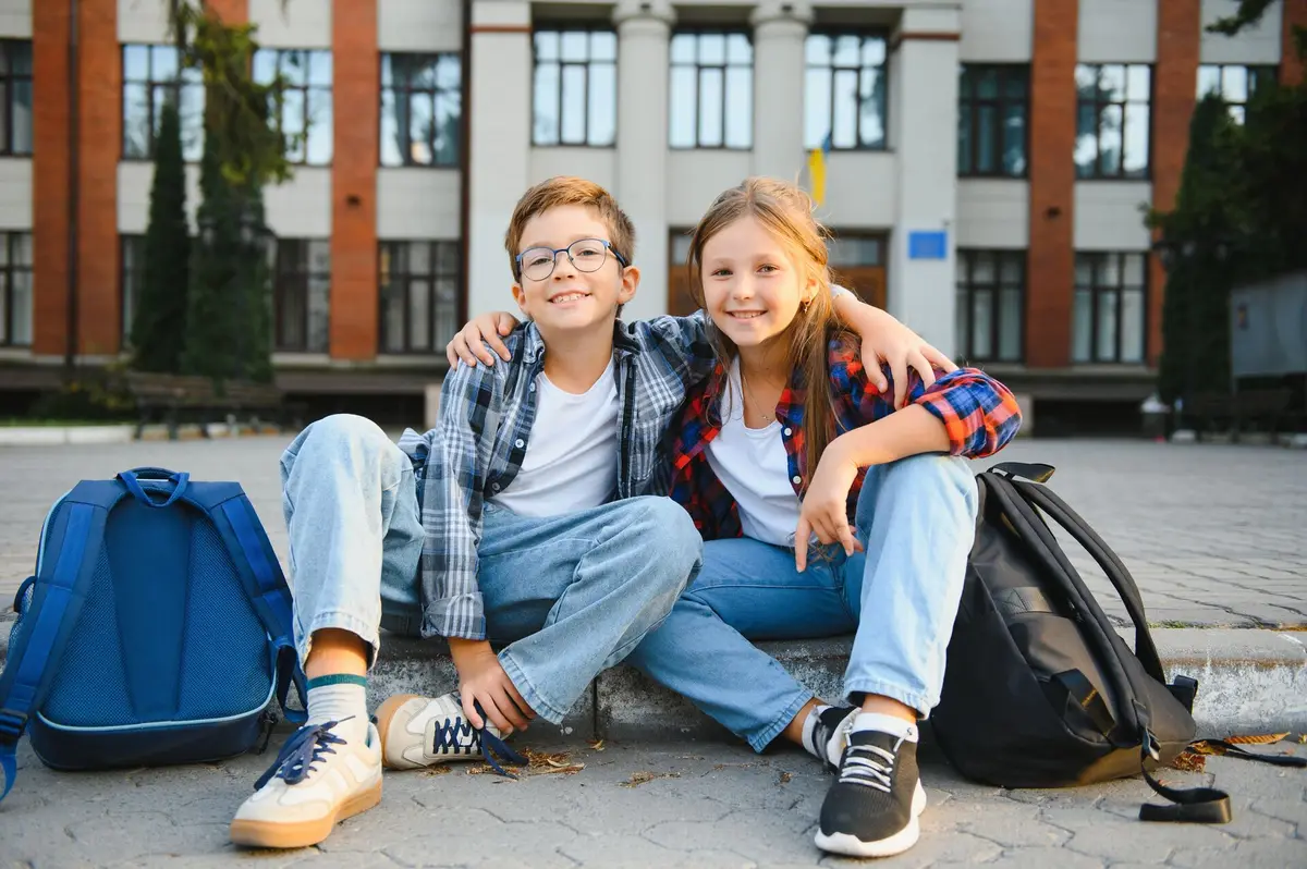 Zwei Kinder sitzen mit ihren Schulranzen vor einem Schulgebäude
