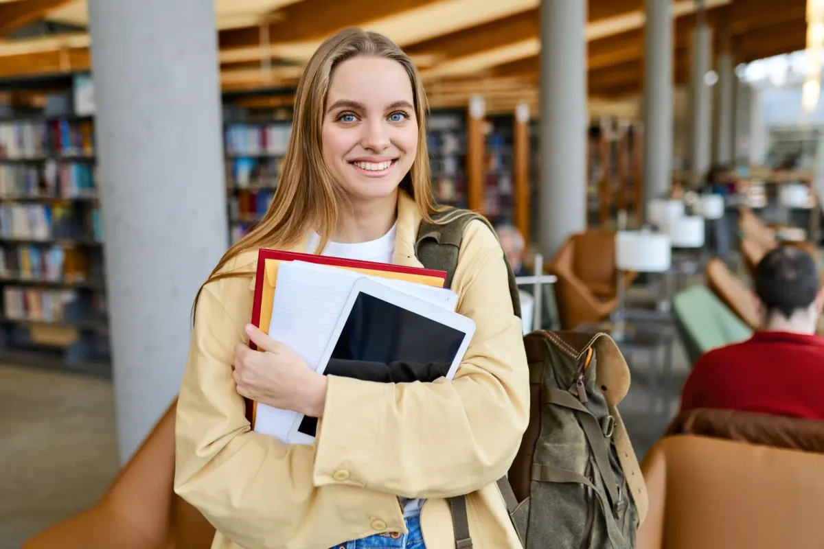 Junge Frau hält Bücher und ein Tablet im Arm und steht in einer Bibliothek