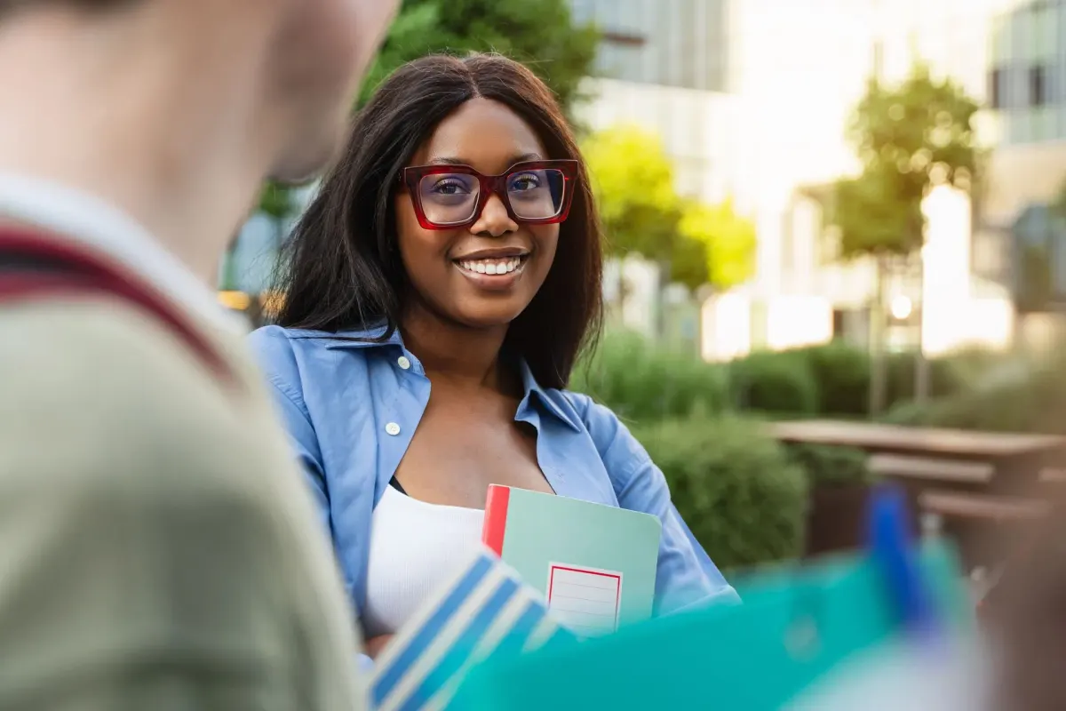 Junge Frau mit Brille hält Bücher im Arm und lächelt, im Hintergrund Grün